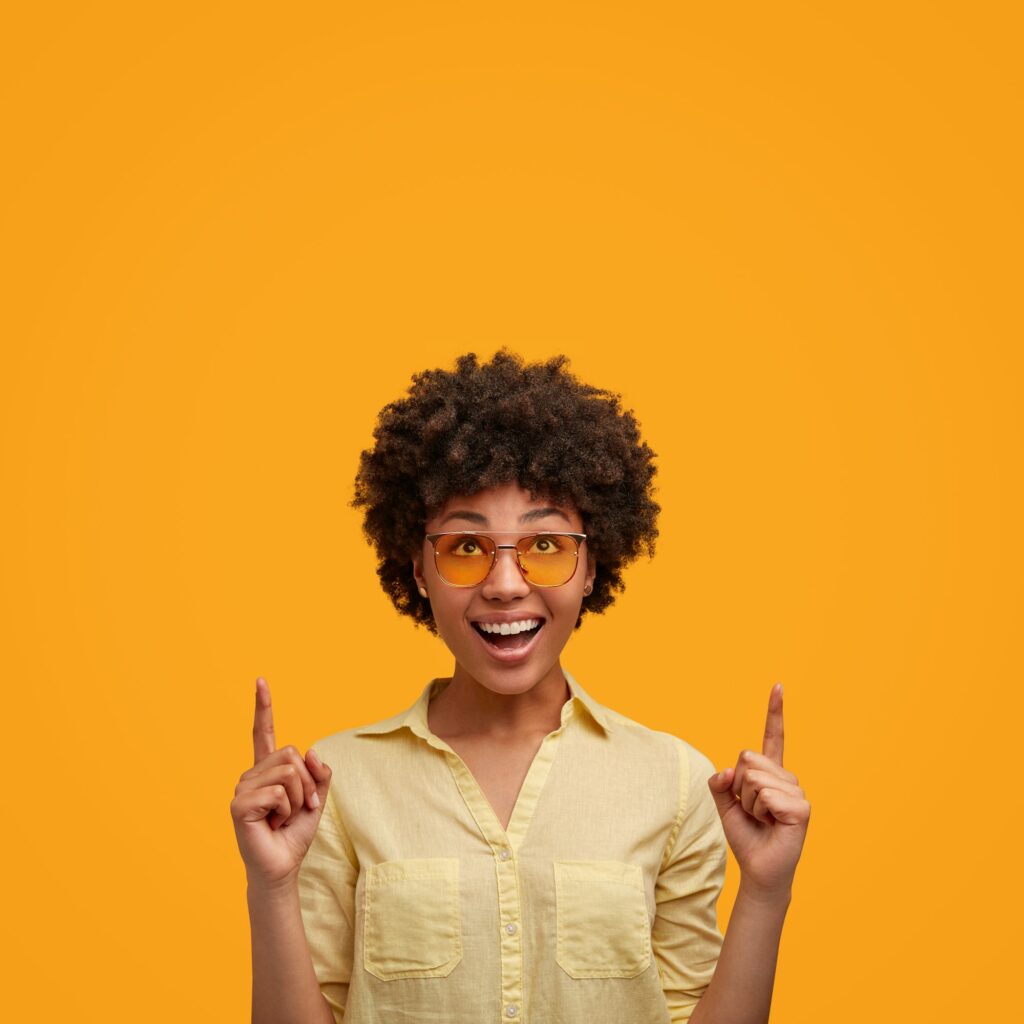 Portrait of glad African American female with joyful expression, keeps fore fingers raised, advertis